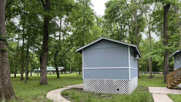 Una piccola baita con base a reticolo bianco e rivestimento grigio, immersa tra alberi e affiancata da un vialetto lastricato.