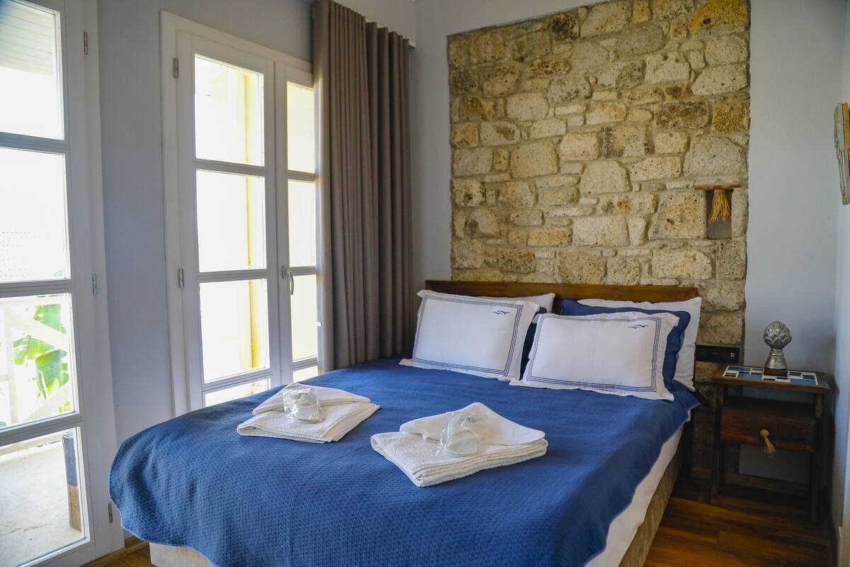 A neatly made bed with a blue bedspread, white pillows, and a wooden headboard. A stone wall and a window with curtains are visible in the background.