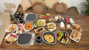 Variedad de alimentos para el desayuno dispuesta sobre una mesa de madera con individuales tejidos, incluyendo huevos, embutidos, frutas y pastelería.