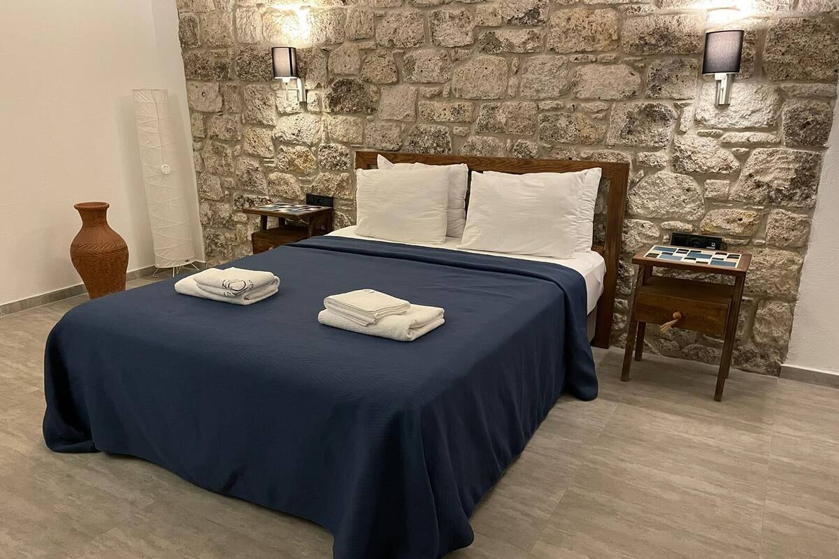 A bed with a dark blue bedspread, white pillows, and two white towels folded on the bed. There are two nightstands with lamps, a wooden side table with a tray, and a stone wall behind the bed.