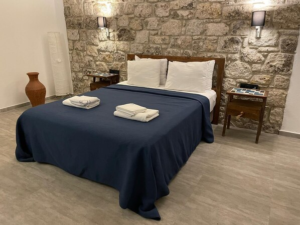 A bed with a dark blue bedspread, white pillows, and two white towels folded on the bed. There are two nightstands with lamps, a wooden side table with a tray, and a stone wall behind the bed. - Adacan Alacatı (Cesme)