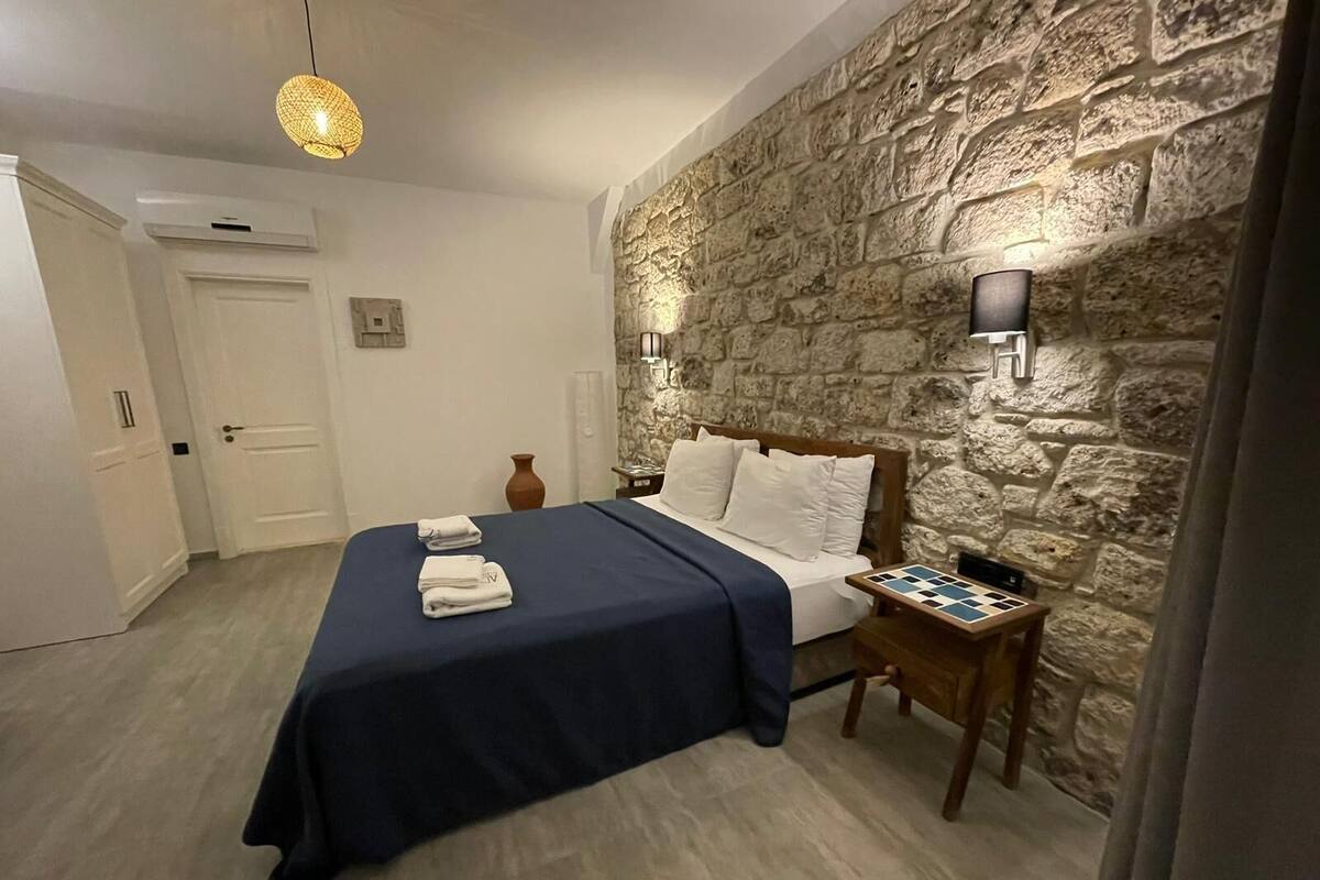 A bedroom with a stone wall, a bed with a blue bedspread, two bedside tables with lamps, and a checkered table.