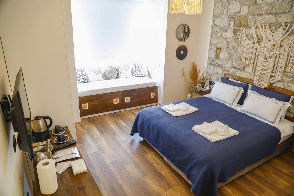 A neatly arranged bedroom with a bed, a wooden dresser, a window with a view, and a stone wall with macramé decorations.