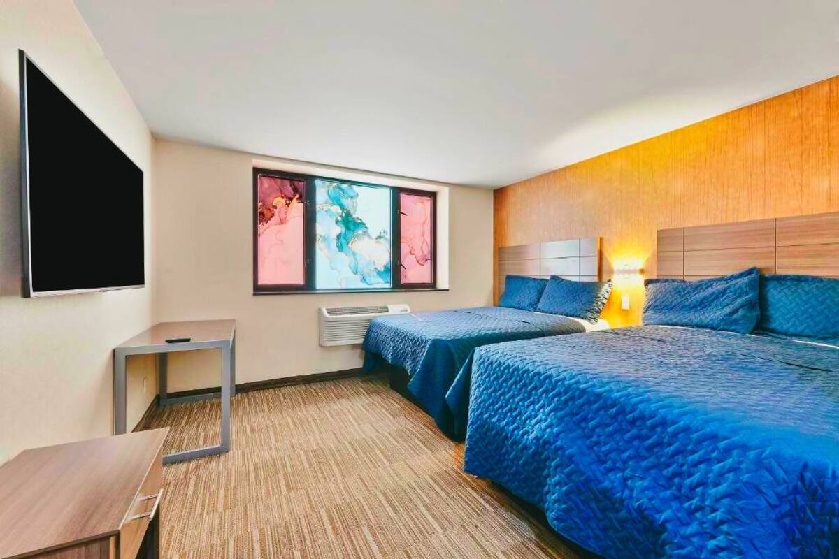 Ein Hotelzimmer mit zwei Betten, einem Flachbildfernseher, einem kleinen Tisch und einem Fenster mit abstrakter Kunst.