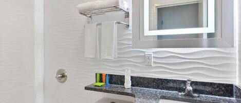 Ein Badezimmer mit weiß gefliesten Wänden, einer Granit-Arbeitsplatte, einem Waschbecken, einem Spiegel und einem Handtuchhalter.