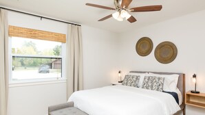 A bedroom with a bed, a bench, a ceiling fan, a window with curtains, and two wall-mounted decorative plates.
