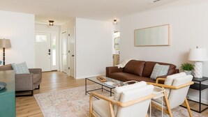 A modern living room with a brown leather sofa, a glass coffee table, and a beige armchair. There is a white door leading to another room, and a mirror on the wall.