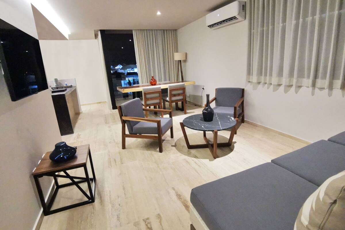 Un salon moderne comprenant un canapé, deux fauteuils, une table basse et un téléviseur fixé au mur. On y trouve également un coin repas avec une table et des chaises, ainsi qu’un appareil de climatisation fixé au mur.