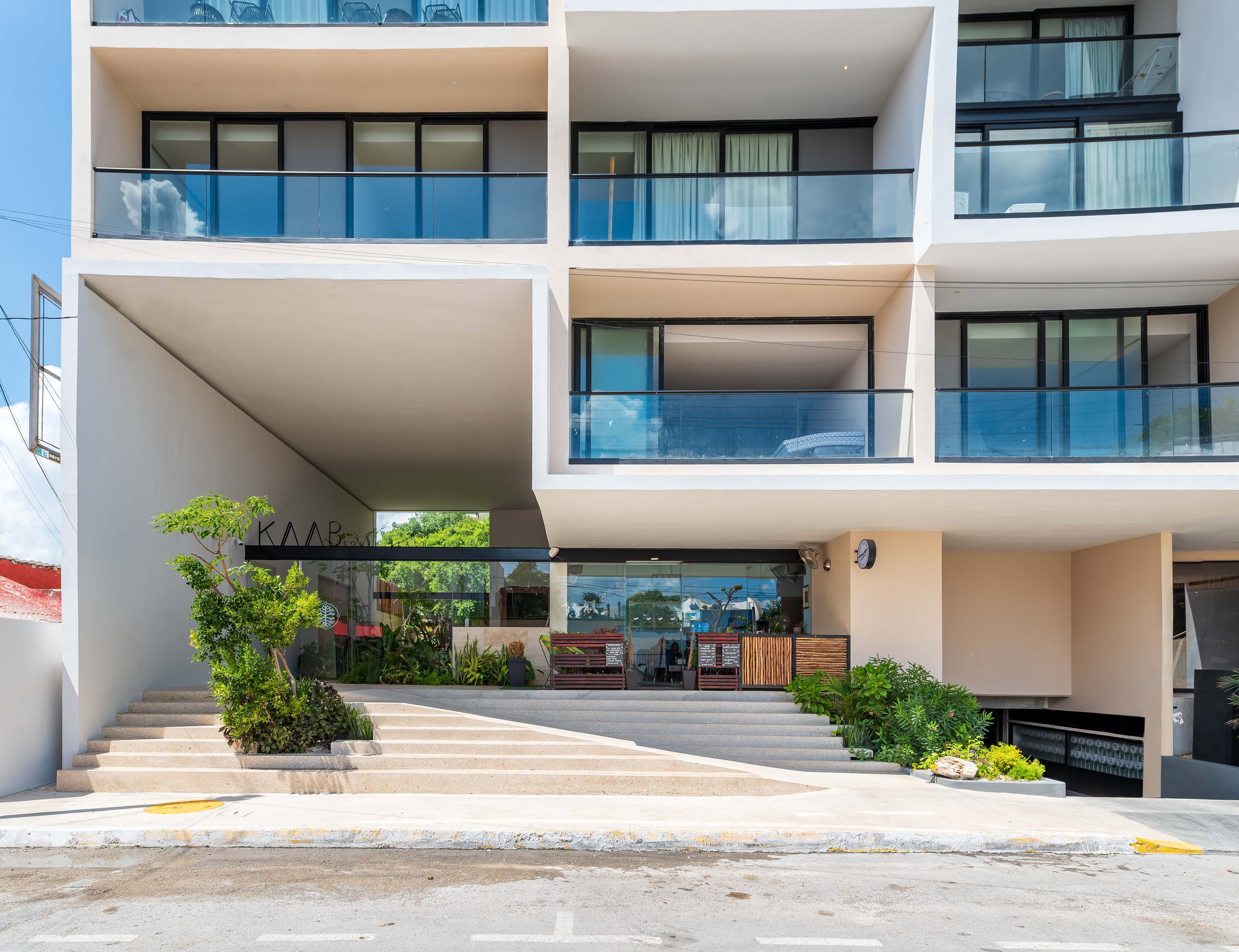 A modern multi-story building with balconies, a staircase leading to the entrance, and a small outdoor seating area with plants.