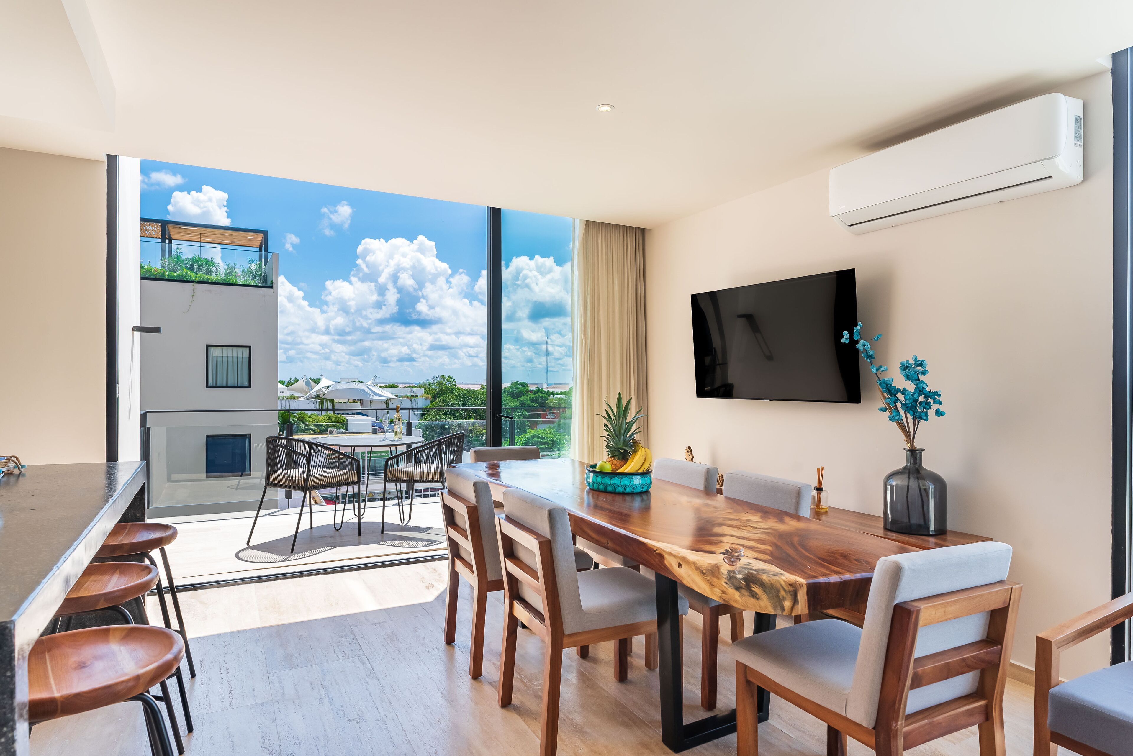 A modern dining area with a wooden table and chairs, a TV mounted on the wall, and a view of the sky and greenery outside.