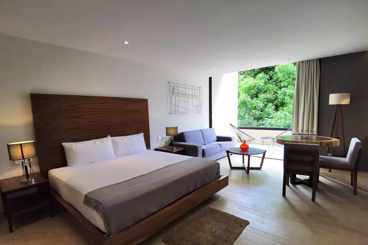 Une chambre d’hôtel moderne dotée d’un grand lit, d’un coin salon avec un canapé et des fauteuils, d’une petite table et offrant une vue sur la verdure environnante.
