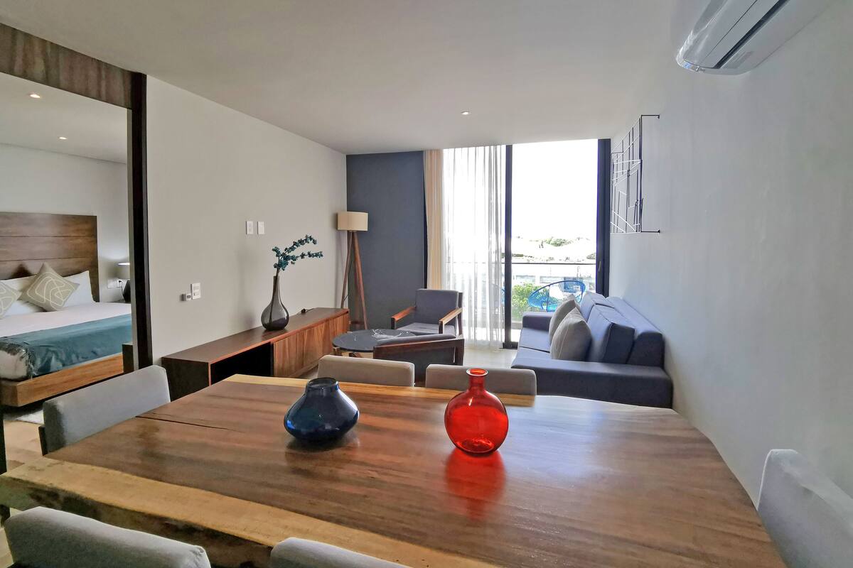 Une chambre d’hôtel moderne avec une table à manger en bois, un canapé, un lit avec une tête de lit et une vue sur l’extérieur grâce à de grandes fenêtres.