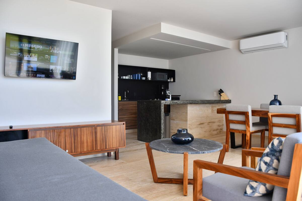 Un salon moderne comprenant un canapé, une table basse et un coin repas avec des chaises et une petite table. Il y a également un coin cuisine équipé d’un four à micro-ondes et de placards.