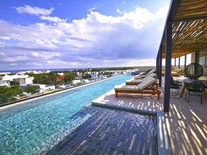 Une piscine avec des chaises longues et des tables, une vue dégagée sur l’océan et un cadre tropical, avec des bâtiments en arrière-plan.