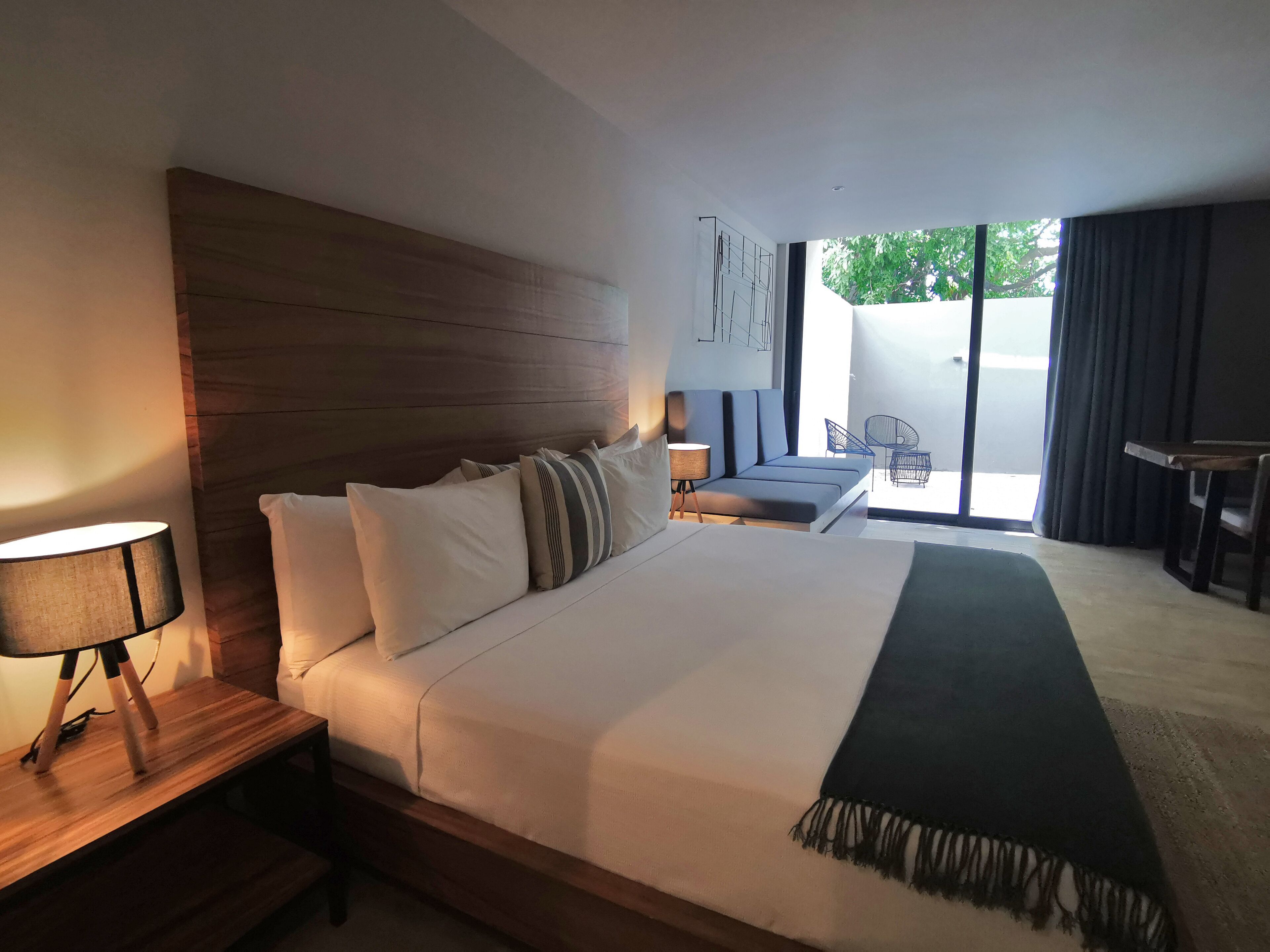 A modern hotel room with a large bed, a wooden headboard, a bedside table with a lamp, a small seating area with a sofa and chairs, and a view of the outdoors through a large window.