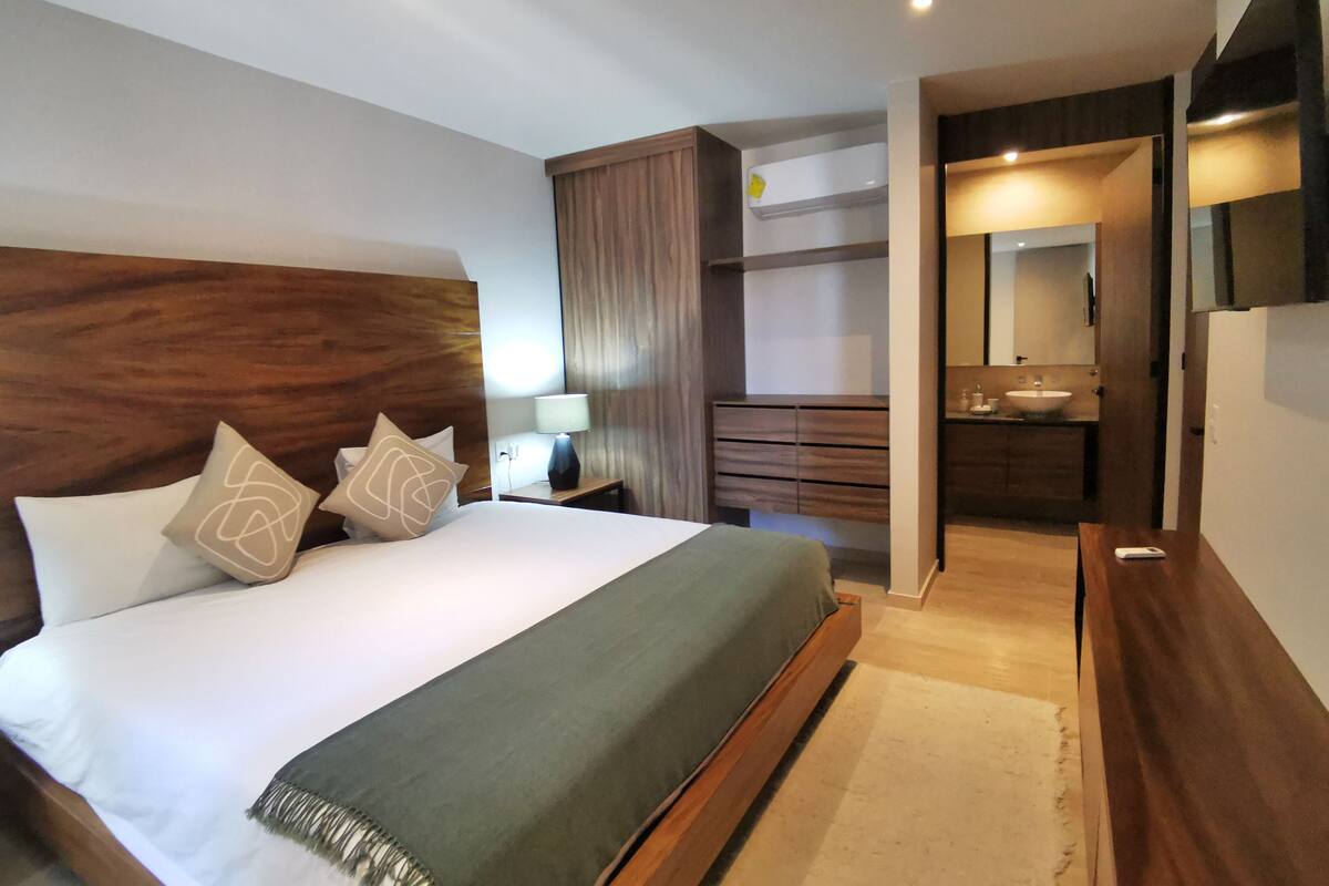 Une chambre d’hôtel moderne avec un grand lit, une tête de lit en bois, une table de chevet avec une lampe, une armoire, une salle de bain avec un lavabo et un miroir, et un téléviseur à écran plat fixé au mur.