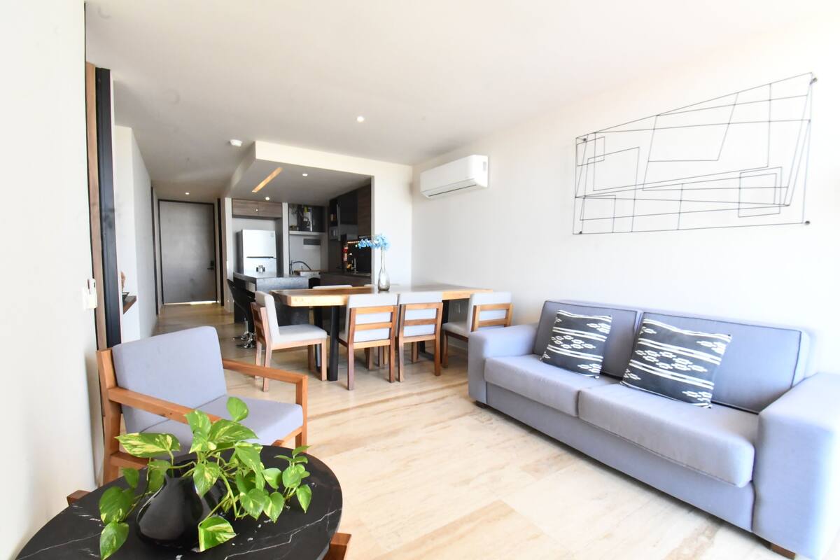 Un salon moderne avec un canapé gris, un coin repas avec des chaises, une table noire agrémentée d’une plante, et une cuisine en arrière-plan.