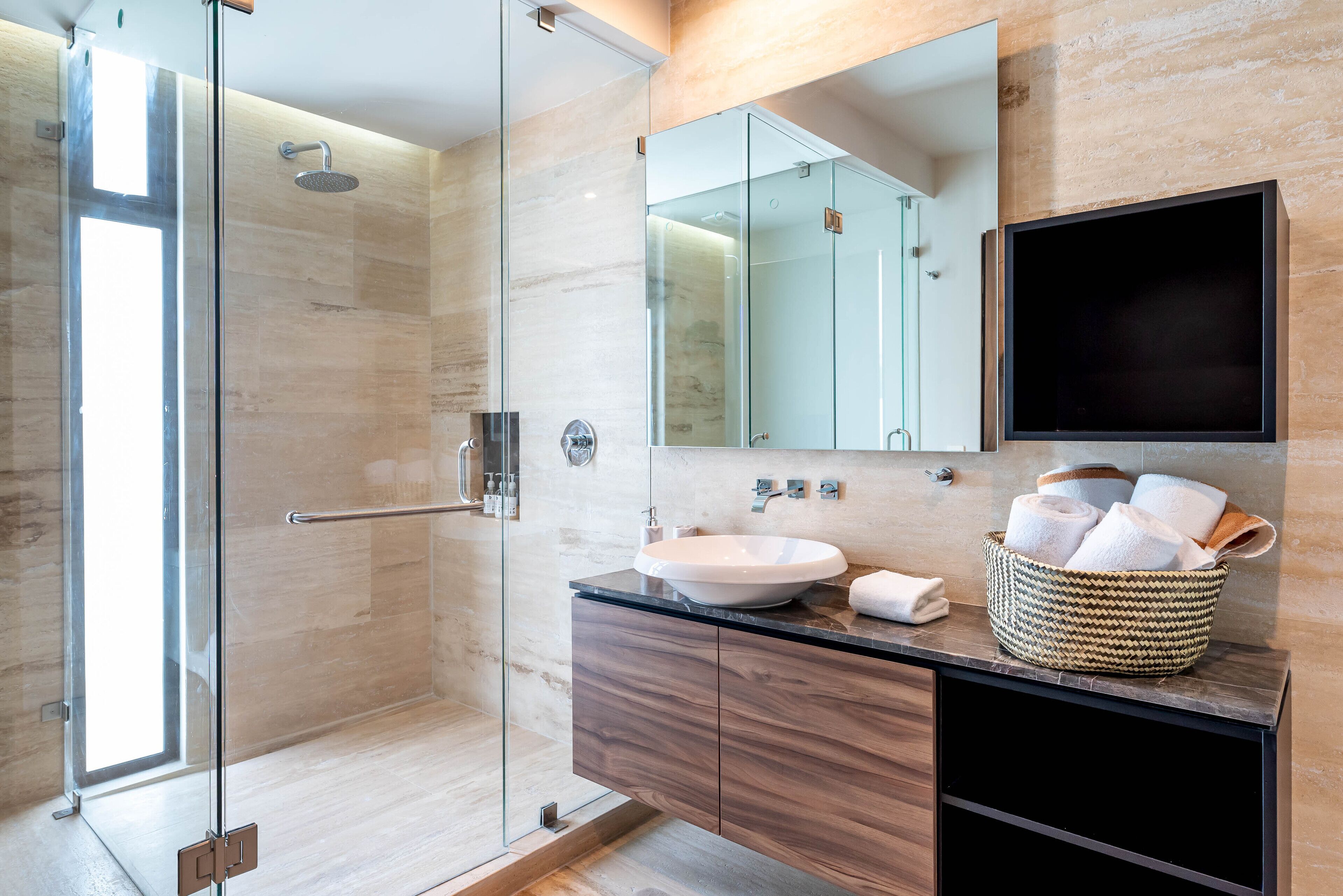 Un baño moderno con ducha de vidrio, un espejo grande y un tocador de madera con lavamanos y cesta para toallas.