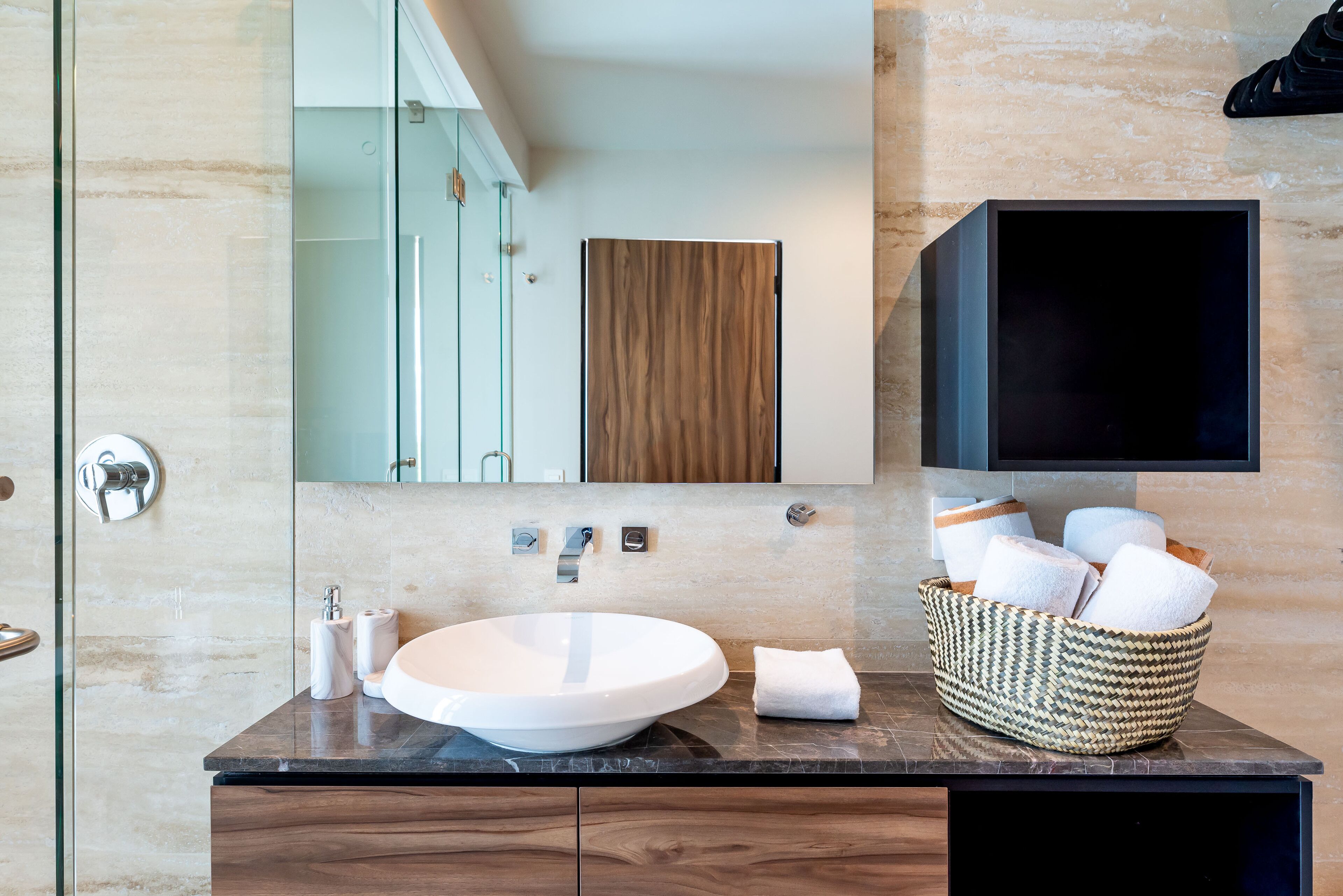 Un baño moderno con un lavamanos blanco redondo, un mueble de tocador de madera, un espejo grande y una cesta tejida con toallas.