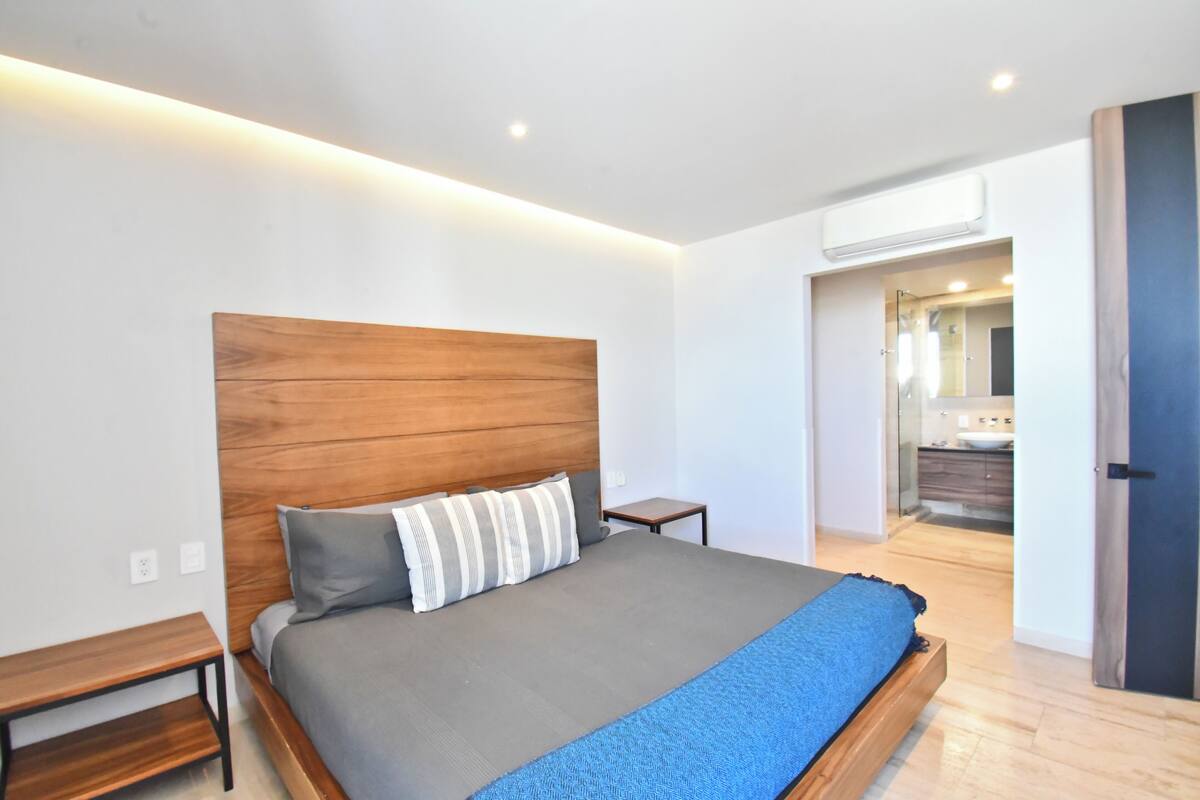 Une chambre moderne avec une tête de lit en bois, une couvre-lit grise, une couverture bleue, une table de chevet et une petite table de nuit.