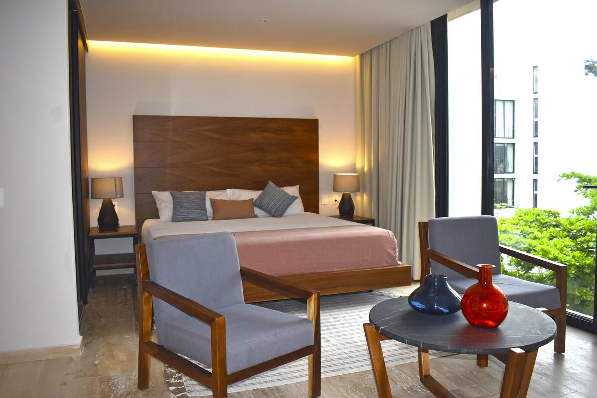 Une chambre d’hôtel avec un grand lit, deux fauteuils, une petite table ornée de vases et une vue sur la verdure environnante.