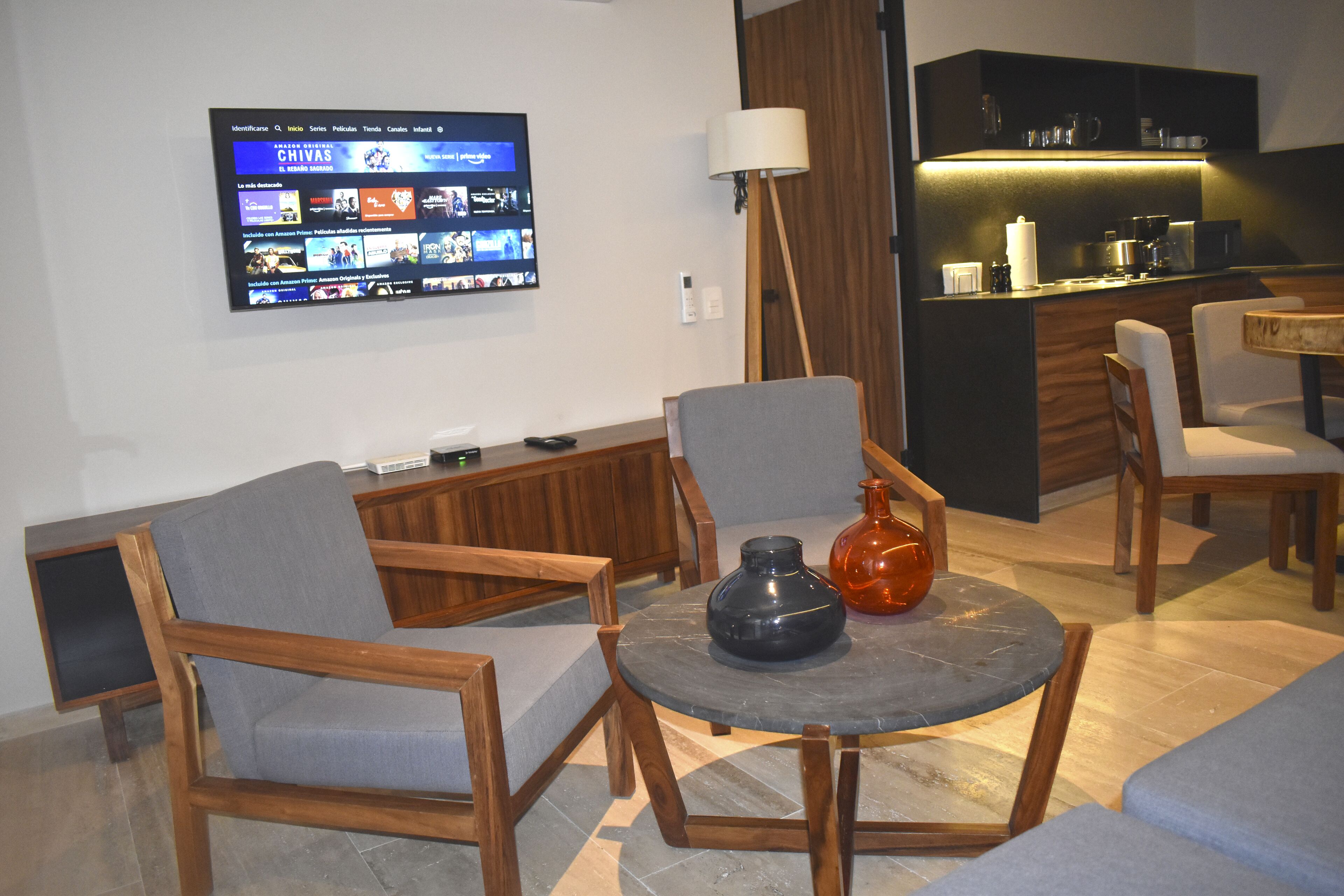 A modern living room with wooden furniture, a flat-screen TV mounted on the wall, and a kitchen area in the background.