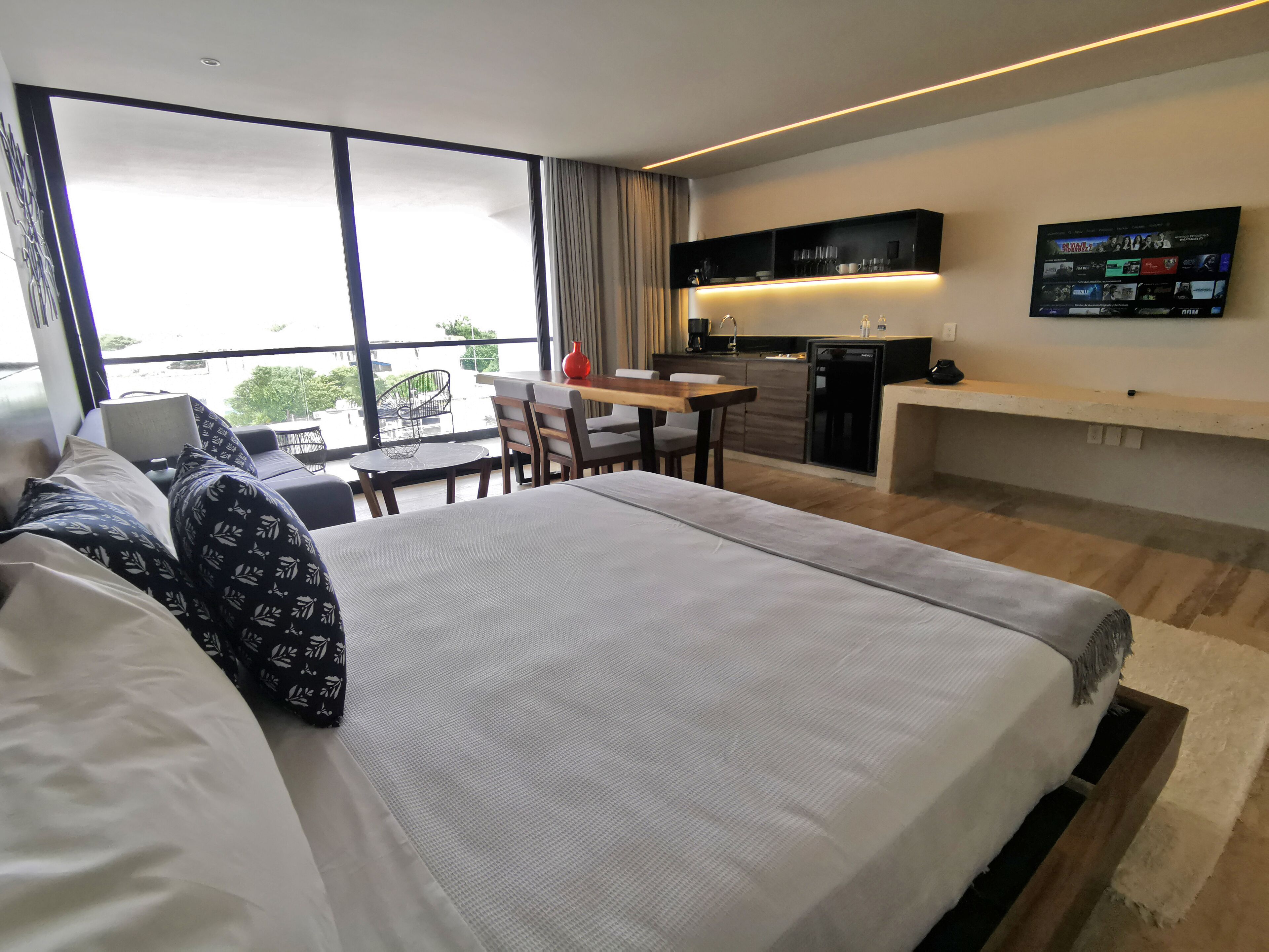 A modern hotel room with a large bed, a dining area, a kitchen, and a TV mounted on the wall.