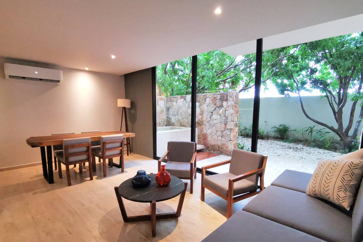 Un salon moderne comprenant un canapé, des fauteuils, une table à manger et une table basse. On y trouve un mur en pierre et une vue sur les arbres à l’extérieur.