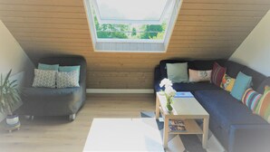 Ein gemütliches Zimmer mit schrägen Wänden, einem Oberlicht, einer Couch, einem Sessel und einem kleinen Tisch mit Blumenvase.