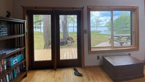 A room with a wooden floor, a bookshelf, a sliding glass door leading to an outdoor deck, and a window with a view of a lake and trees.