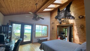 A bedroom with a bed, a desk with a computer, a ceiling fan, and a large window overlooking a lake.