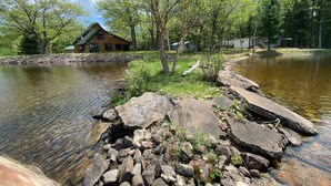 A cabin by a lake with a stone path leading to it, surrounded by trees and a clear sky.