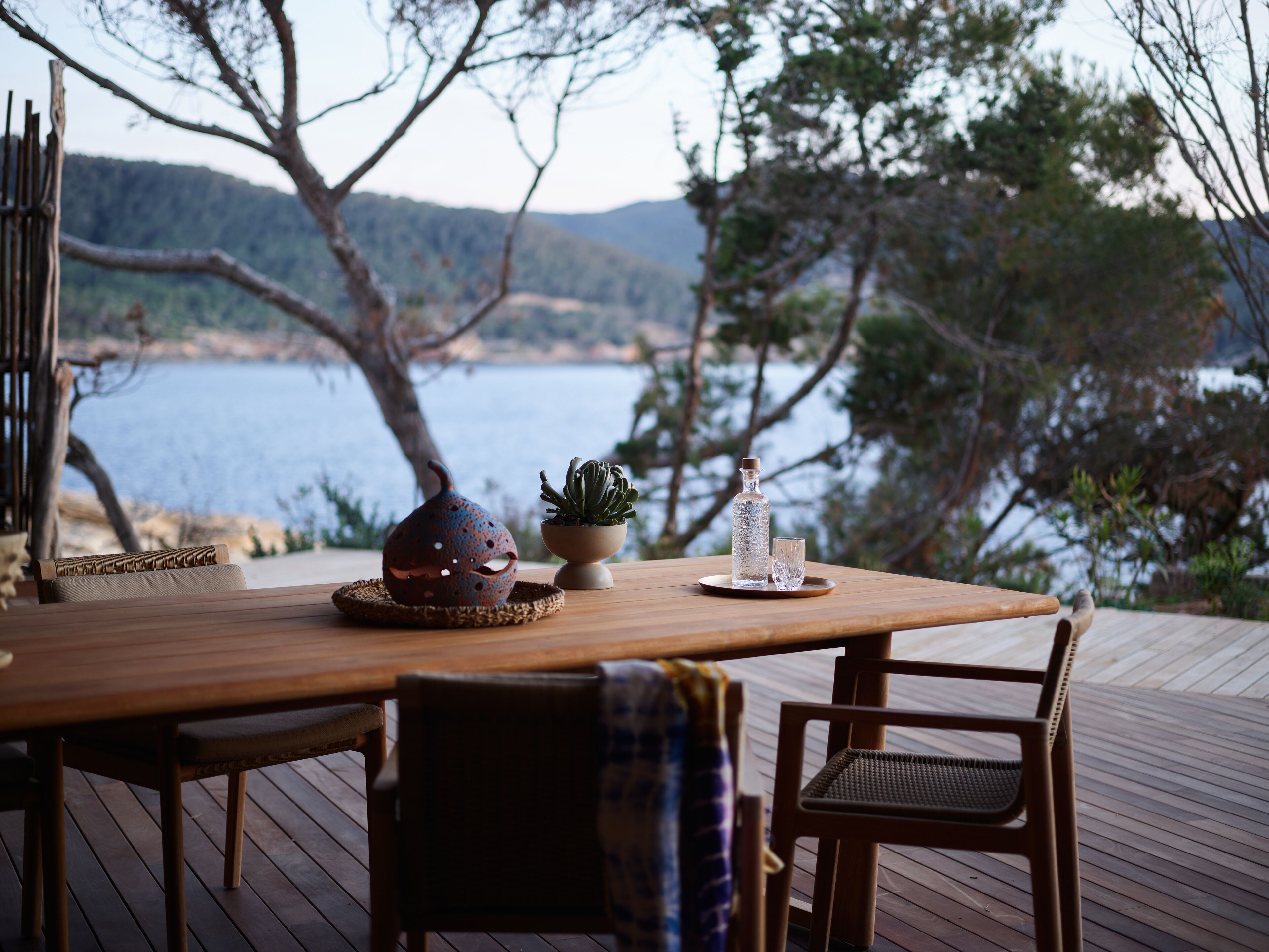 A wooden deck with a table set for two, a chair, and a view of a lake and hills in the background.