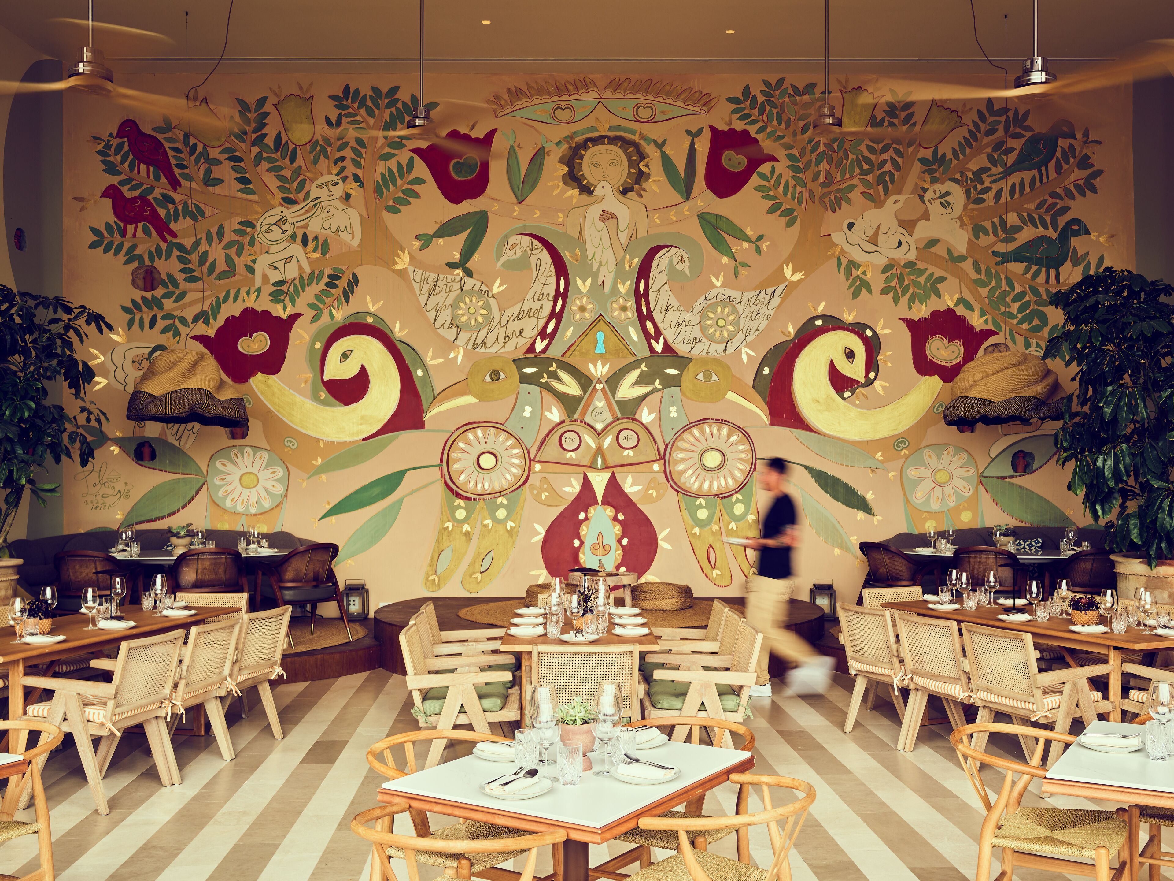 A restaurant with wooden tables and chairs, a large mural on the wall, and potted plants.