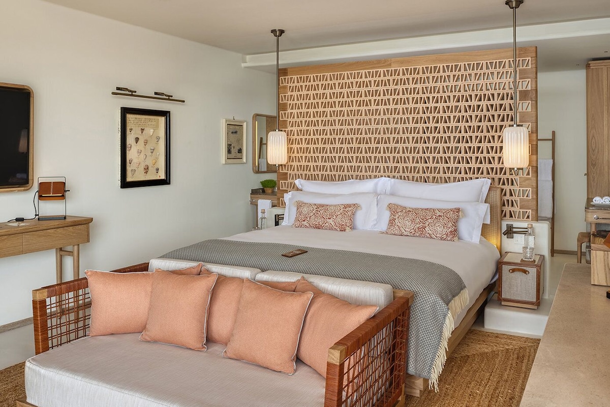 A modern hotel room with a large bed, a wooden headboard, a desk, and a wicker sofa.