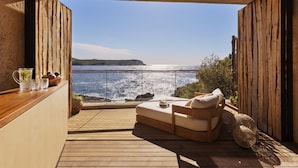 A balcony with a wooden floor, a cushioned seating area with a hat and a bowl, and a view of the sea. - Six Senses Ibiza (Sant Joan de Labritja)