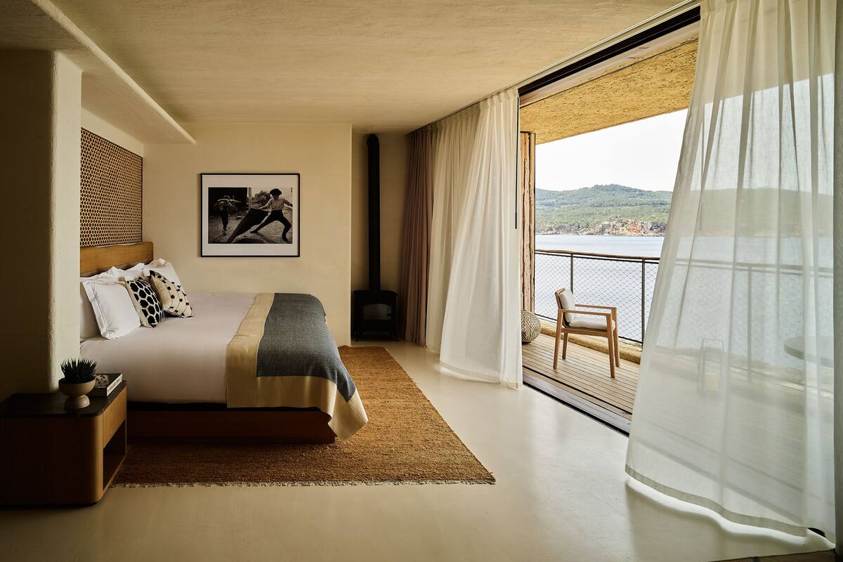 Een slaapkamer met een bed, nachtkastje, stoel, groot raam met uitzicht op zee en een lijst met een afbeelding aan de muur.
