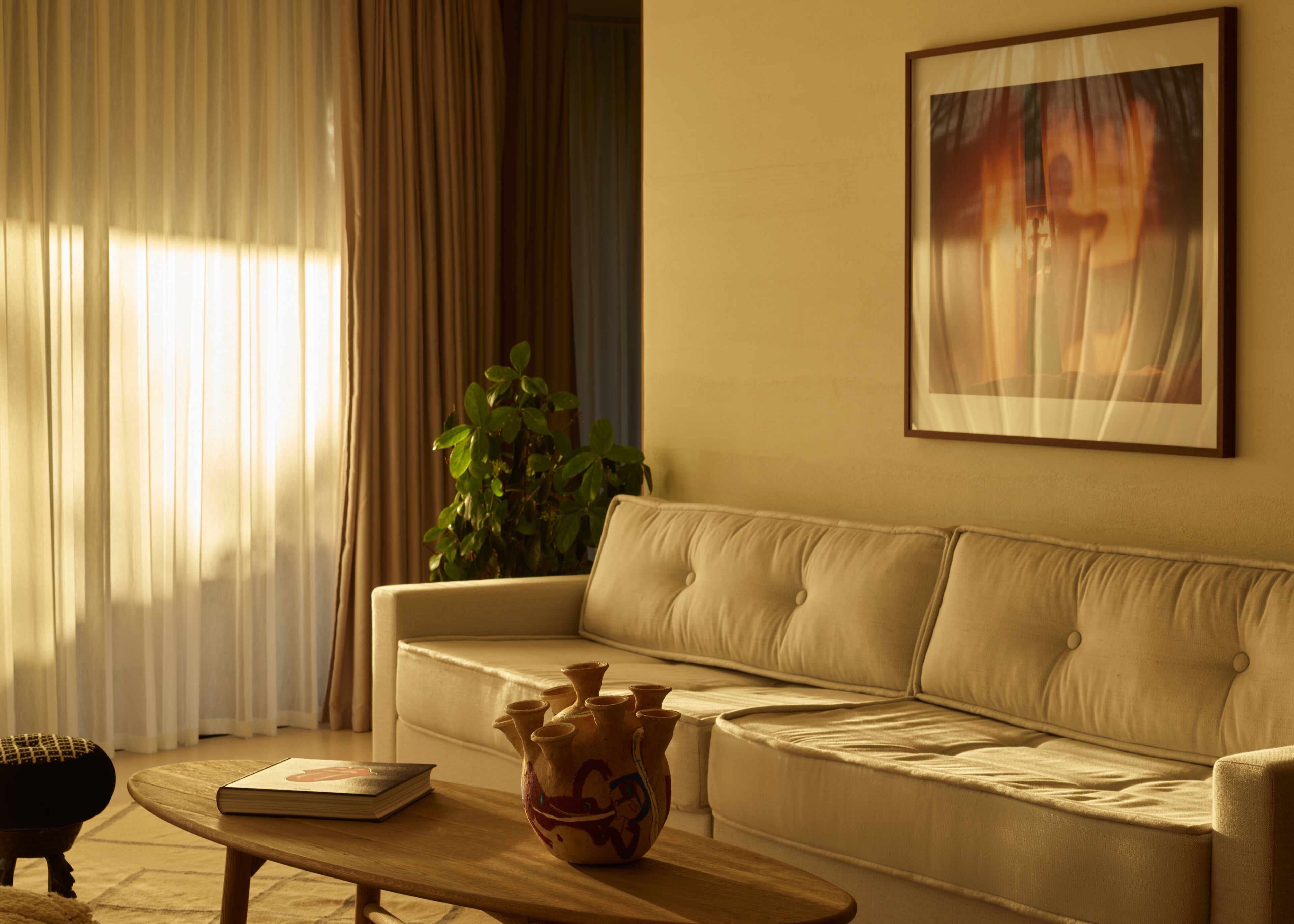 A living room with a beige sofa, a coffee table with a book and a decorative vase, a potted plant, and a painting on the wall.