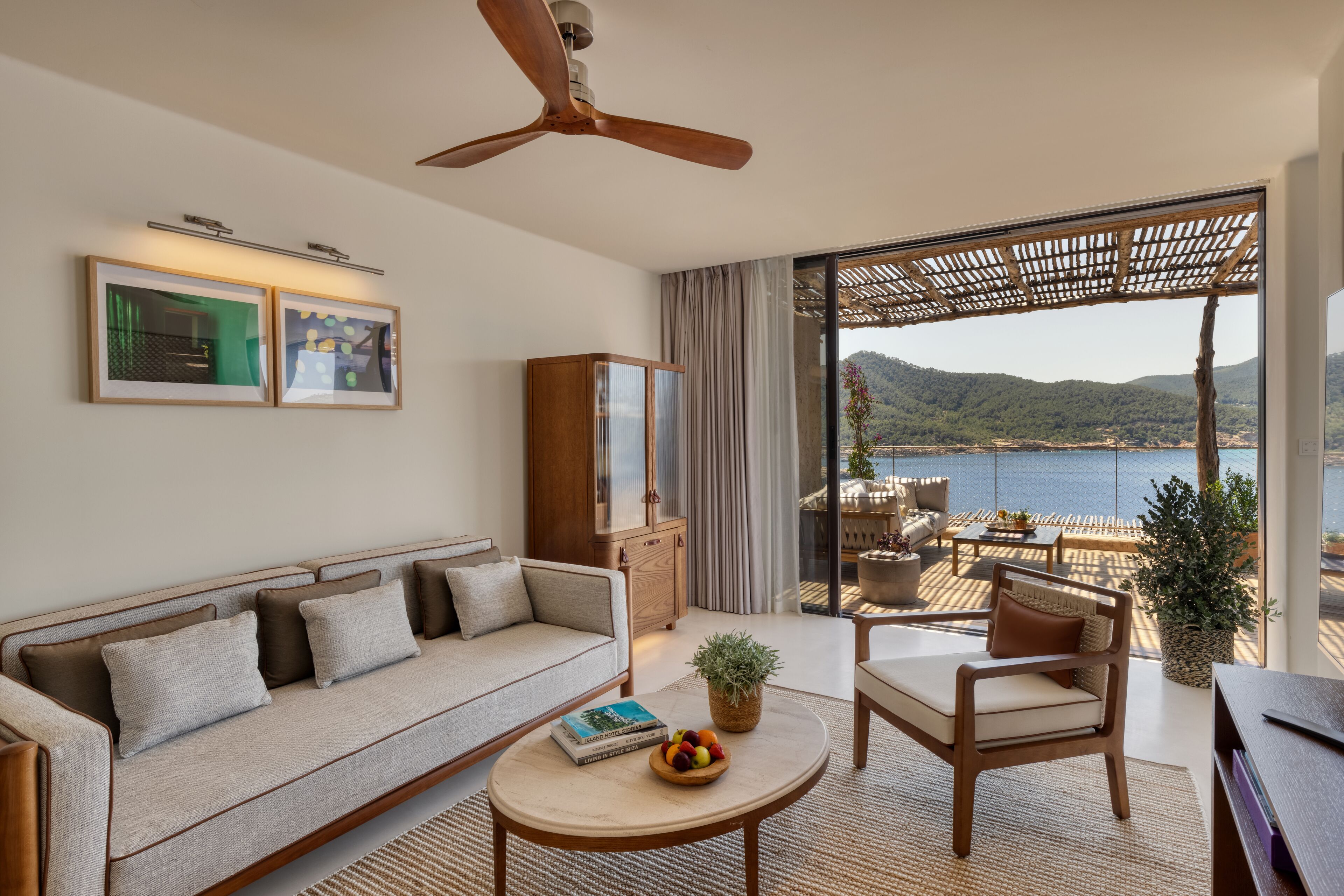 Una sala de estar moderna con sofá, sillón y mesa de centro, un ventilador de techo y vistas a un cuerpo de agua y montañas a través de amplios ventanales.