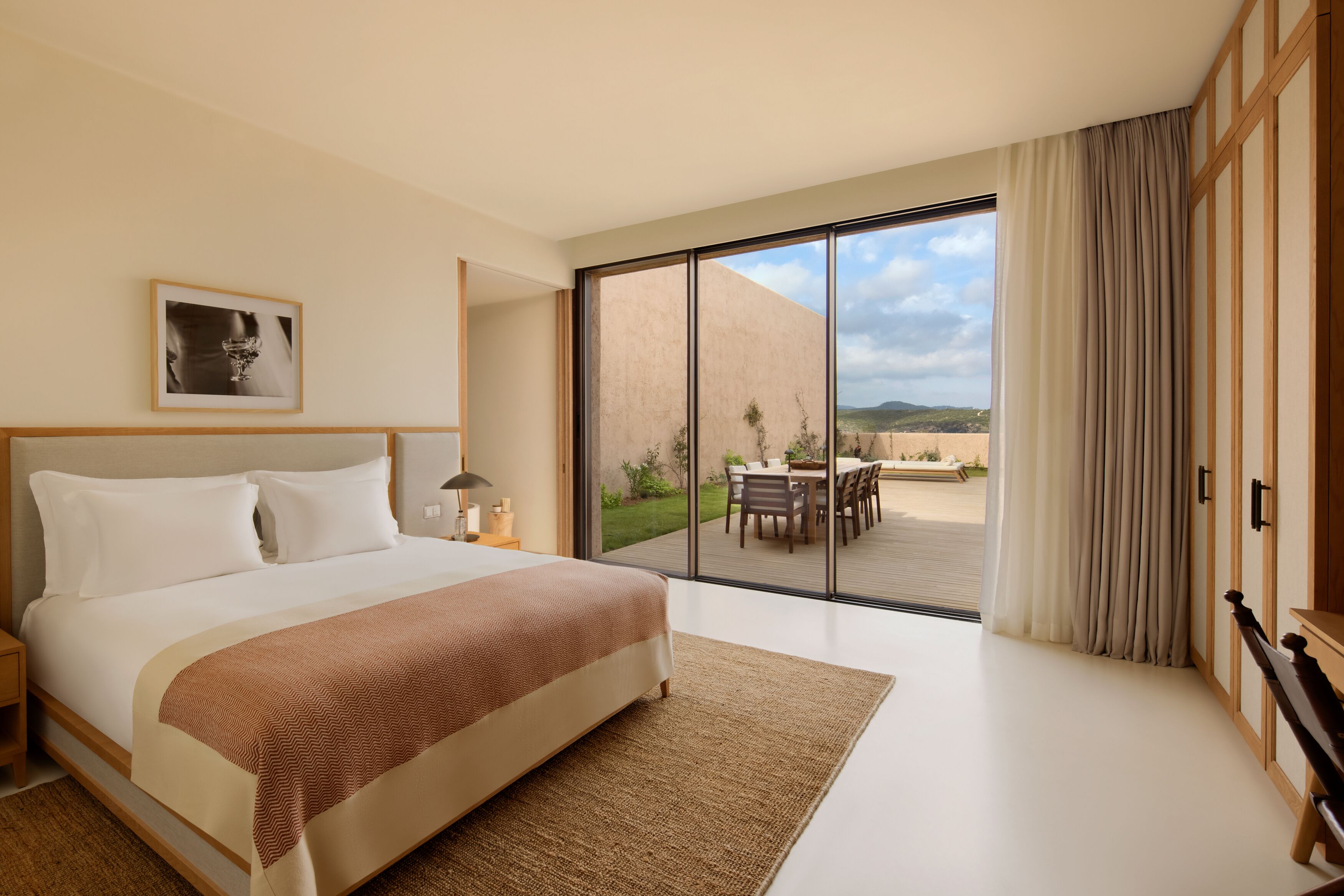 A modern hotel room with a large bed, a view of the outdoors, and a dining area visible through the glass door.