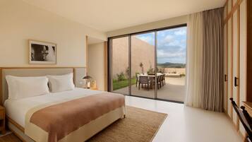 A modern hotel room with a large bed, a view of the outdoors, and a dining area visible through the glass door.