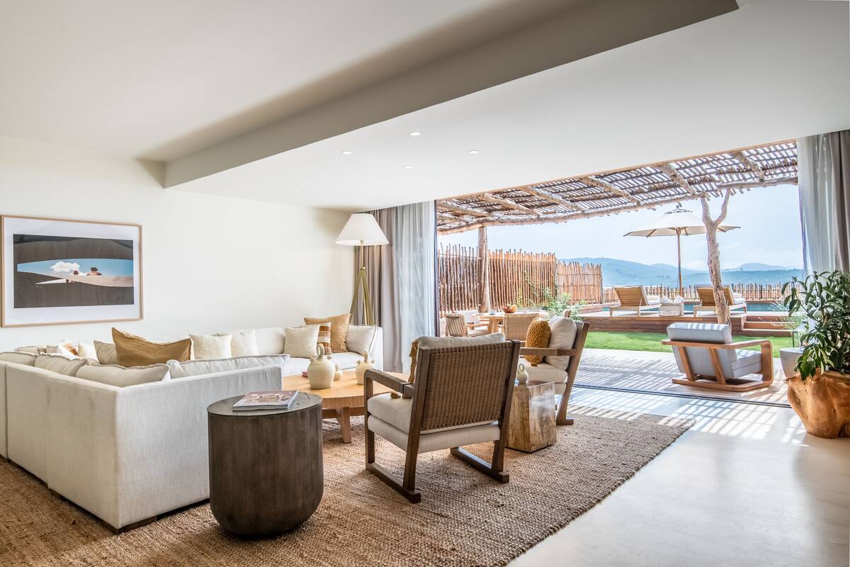 Een ruime woonkamer met een grote bank, een houten stoel en een salontafel. Er is uitzicht op een terras met lounge fauteuils en een zwembad.