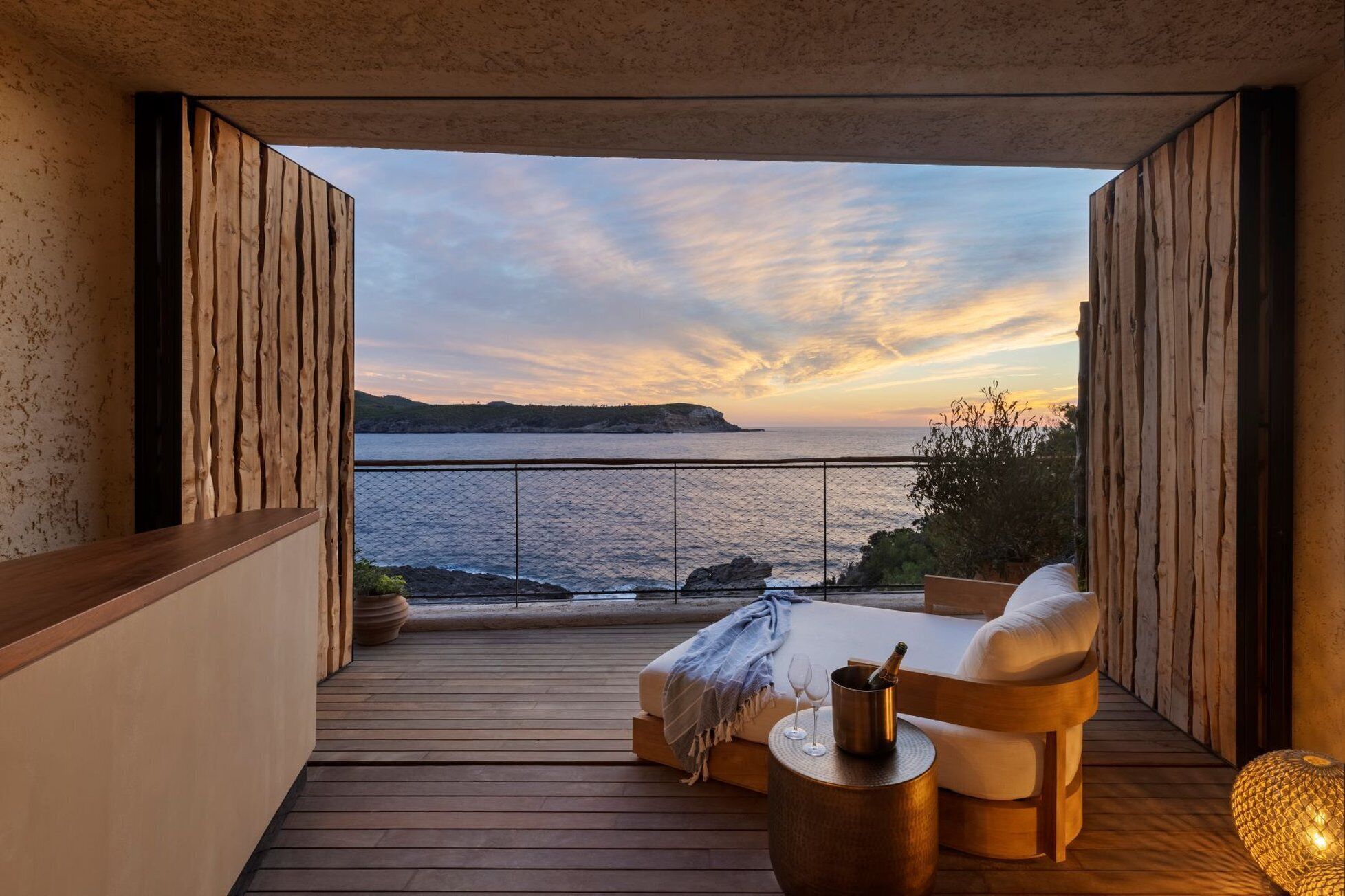Un balcón con suelo de madera, una zona de estar con cojines y vista al mar al atardecer.