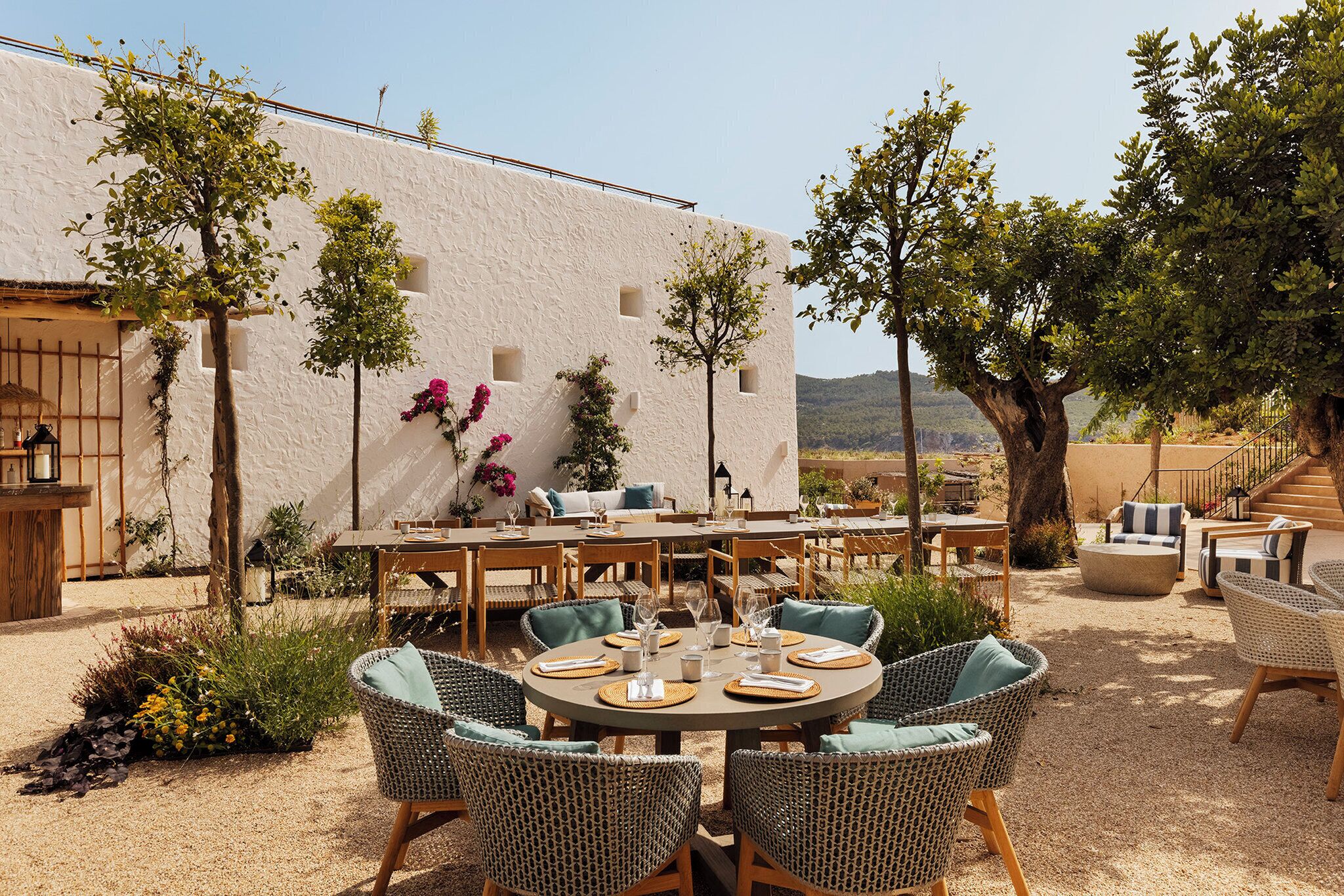 An outdoor patio with round tables and wicker chairs, surrounded by a white building with windows and a view of hills.