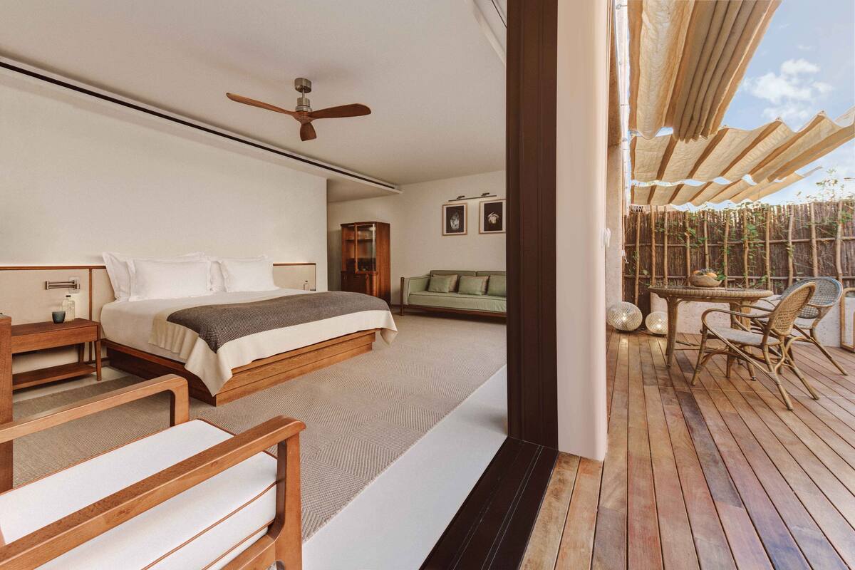 Een slaapkamer met een groot bed, een nachtkastje, een houten bankje en uitzicht op een balkon met rieten meubels en een tafel.