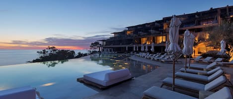An infinity pool with lounge chairs and umbrellas, overlooking a sunset.
