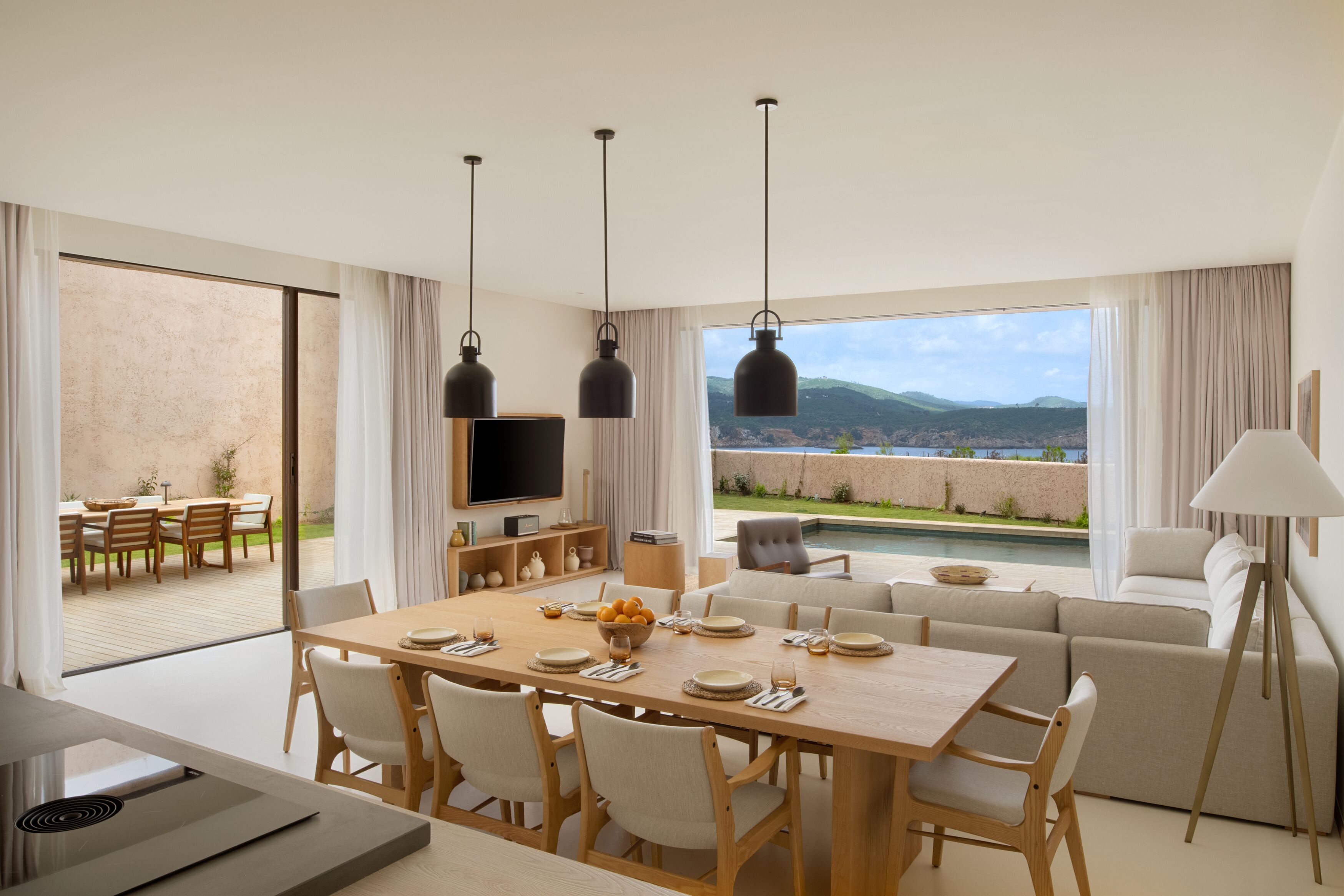 Un comedor moderno con una mesa grande, sillas y un televisor. Se ve un sofá y una lámpara al fondo.