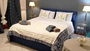 Una cama bien hecha con cabecera azul y ropa de cama blanca, dos lámparas de noche, una mesita redonda con un jarrón y una silla azul.