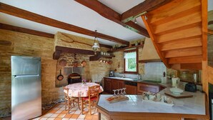 A rustic kitchen with a tiled floor, wooden beams, a refrigerator, a dining table with chairs, and a window with curtains. - Charme et Tradition (trefumel)