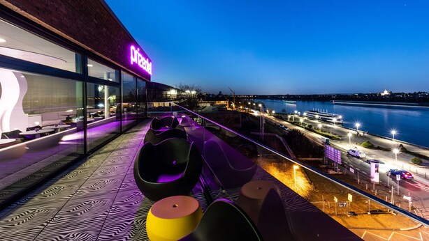 A modern building with a 'Pizza' sign, outdoor seating with unique black and yellow chairs, and a view of a body of water and city lights at dusk. - Prize by Radisson, Bonn City (Bonn)